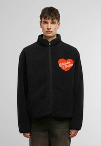 Black fleece jacket with a high collar and full zip. Features an orange heart logo with "Market Studios" text on the left chest.