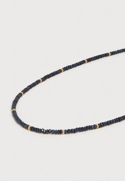 Λεπτό κολιέ με μικρές σκουρομπλε πολύγωνες χάντρες διακοσμημένες με ομοιόμορφα τοποθετημένες χρυσές χάντρες σε ανοιχτό φόντο.