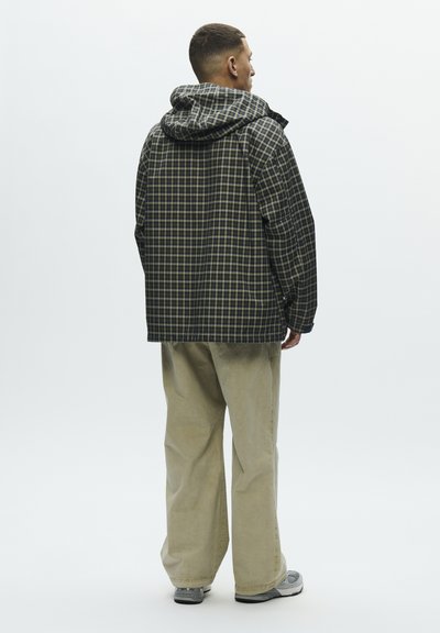 Uomo in piedi di spalle, che indossa una giacca a quadri scura con cappuccio, pantaloni beige a gamba larga e scarpe da ginnastica grigie su uno sfondo semplice.