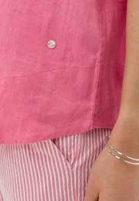 Ingrandimento di una persona che indossa una blusa rosa con un bottone e pantaloni a righe rosa e bianchi, mostrando il braccio con braccialetti argentati.