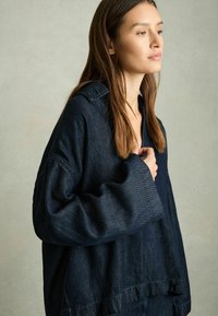 Dunkelblaue Jeanshemd mit lockerer Passform, großem Kragen und weiten Ärmeln. Verfügt über einen ausgefransten Saum und minimale Nähte.