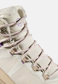 Beżowy but sportowy z teksturowanej tkaniny, syntetycznymi nakładkami, fioletowymi sznurówkami w kropki oraz wyściełanym kołnierzem; charakteryzuje się unikalnym wzorem podeszwy.