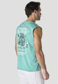 Hombre con camiseta sin mangas color azul verdoso con diseño de dragón japonés blanco y texto en la espalda, combinado con pantalones blancos.