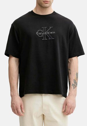 Uomo con una t-shirt nera Calvin Klein con logo e pantaloni bianchi, che mostra un tatuaggio sull'avambraccio con disegni floreali e di pugnale.