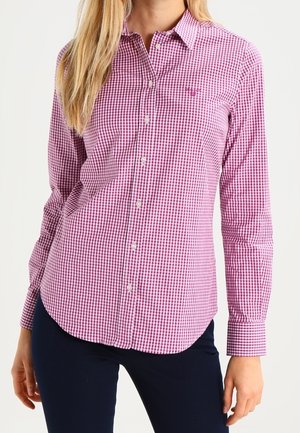 Femme portant une chemise à boutons à carreaux rose et blanc à manches longues et un pantalon bleu marine, debout devant un fond uni.