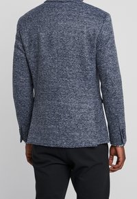 Marinblå strukturerad blazer med ett subtilt mönster, slagkrage och knäppta manschetter, tillsammans med svarta byxor. Bakifrån visas den skräddarsydda passformen.