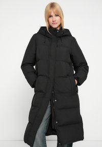 Must pufferjakk, millel on pikk disain, tikk-kangas, kapuuts paeltega ja küljetaskud. Näha on trukid ja tõmblukud.