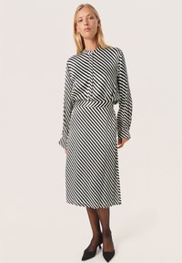 Vrouw in een zwart-witte schuin gestreepte blouse met lange mouwen en bijpassende midi-rok, met zwarte doorschijnende panty's en zwarte hakken.