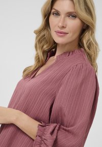 Blusa a maniche lunghe color malva con tessuto testurizzato, scollatura henley e polsini arricciati. Vestibilità comoda con sottili strisce verticali.