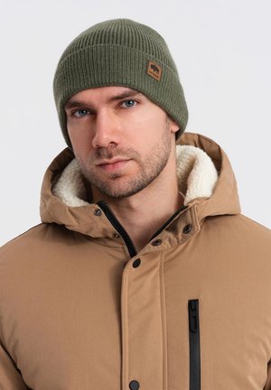 Green knitted beanie with a folded brim and a small brown logo patch, worn with a tan puffer jacket featuring a plush collar and zip.