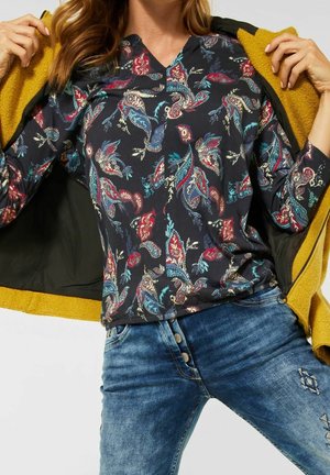 Kobieta ubrana w niebieską, kwiecistą bluzkę z długim rękawem, niebieskie dżinsy z naszywkami, trzymająca otwartą musztardowo-żółtą kurtkę z czarną podszewką.