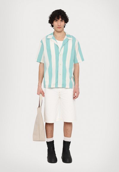 Kortærmet skjorte med knapper i teal- og hvide striber, parret med hvide shorts og bærende en beige taske. Sorte støvler fuldender looket.