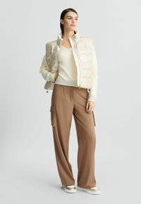 Gilet imbottito beige scintillante, maglione crema a maniche lunghe, pantaloni larghi marroni con tasche cargo e sneakers con dettagli dorati.