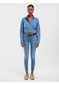 Naine seisab helesinises teksasärgis ja teksapükstes, pruun vöö ja beežide tossudega valge tausta ees.