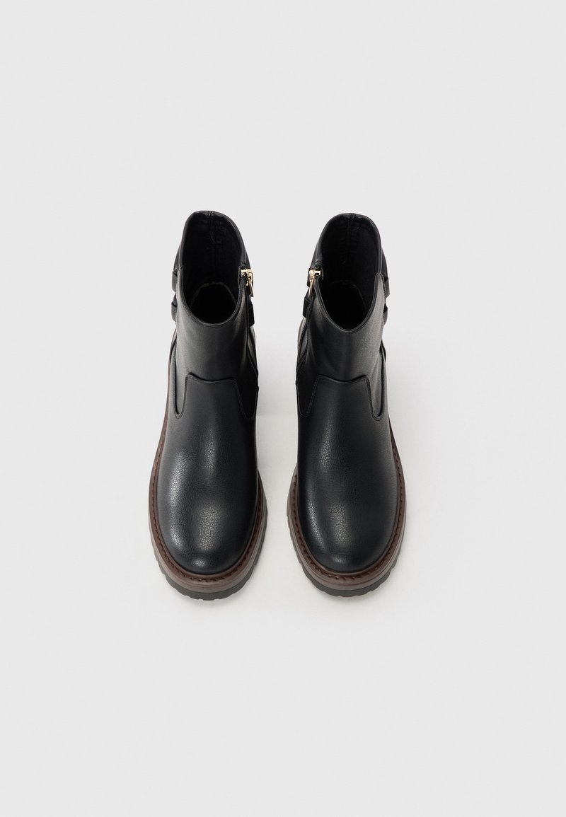 Ein Paar schwarze Lederstiefeletten mit braunen Sohlen und seitlichen Reißverschlüssen, von oben auf einem hellen Hintergrund betrachtet.