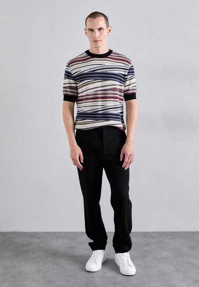 Adīts rakstains apaļkrūšu džemperis bēšā, tumši zila un sarkanā krāsā; kombinēts ar teksturētām melnām biksēm un baltām sporta apaviem. Skaista, ikdienišķa stila.