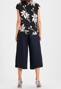 Czarna bluzka z białym kwiatowym wzorem, krótki rękaw, w zestawieniu z granatowymi spodniami culottes. Modelka nosi srebrne szpilki. Widok z tyłu.
