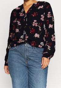 Person som bär en svart långärmad blus med röda och rosa blommönster inskjuten i blå denimjeans.