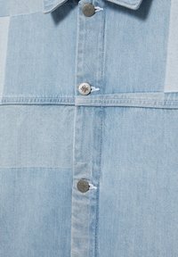 Tissu en denim bleu clair avec un motif patchwork comportant des boutons métalliques argentés avec des motifs gravés le long du centre devant.