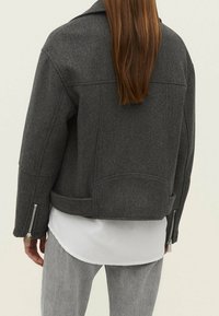 Veste grise en mélange de laine avec un design court, un col pointu et des poignets zippés. Texture lisse avec des coutures nettes et un détail yoke au dos.