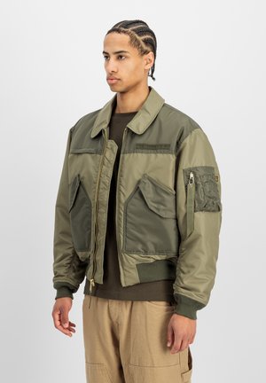 Junger Mann mit geflochtenen Haaren, der eine grün-olive Bomberjacke mit großen Taschen und beige Hosen trägt, steht vor einem weißen Hintergrund.