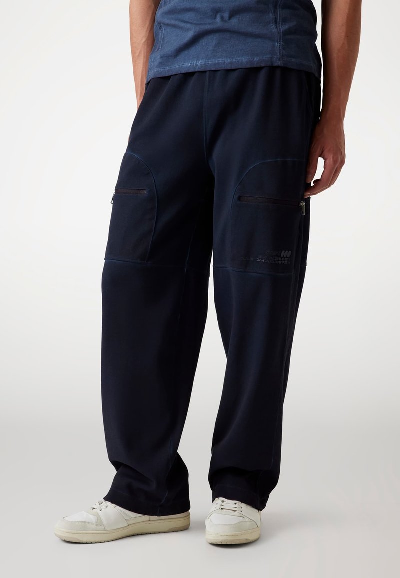 Marineblaue Cargo-Hosen mit Reißverschlüssen an den vorderen Taschen, weitem Bein-Design und glatter Textur, kombiniert mit weißen Sneakers.