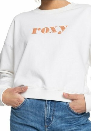 Sweat blanc avec texte orange "roxy", porté par une personne les mains dans les poches d’un jean bleu.