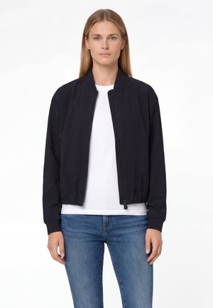 Kobieta ubrana w czarną kurtkę na zamek, białą koszulę i niebieskie dżinsy, stojąca na jednolitym białym tle.