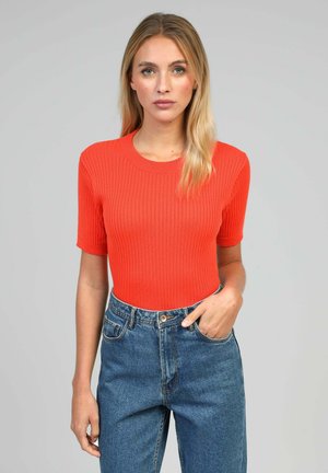 Femme aux longs cheveux blonds portant un haut côtelé orange ajusté à manches courtes et un jean bleu taille haute, main dans la poche, fond neutre.