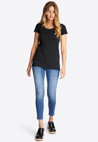 Schwarzes T-Shirt mit kurzen Ärmeln, figurbetontes Design, kombiniert mit hellblauen Skinny-Jeans. Das Model trägt schwarze Clogs mit einer Holzsohle.