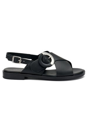 Sandalo in pelle nera con fasce incrociate, elemento circolare, cinturino alla caviglia regolabile, suola piatta e design minimalista.
