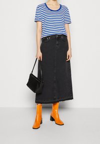 Camiseta a rayas azules y blancas, falda maxi de mezclilla negra, botas de charol naranjas hasta la rodilla y un pequeño bolso negro. Texturas suaves y ajuste relajado.
