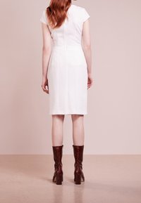 Žena s dlouhými rudými vlasy, oblečená v upnutých bílých šatech a hnědých kozačkách na podpatku do poloviny lýtek, stojící na světle růžovém pozadí.