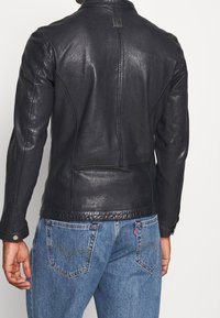 Svart läderjacka med texturerad finish, skräddarsydd passform och långa ärmar. Har subtila metallaccenter och sydda detaljer på baksidan.