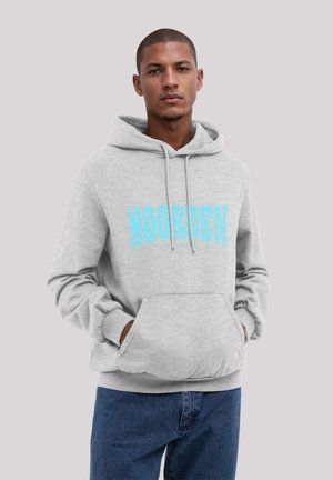 Young man with short hair wearing a light gray hoodie with blue "NOORDEN" text, hands in pocket, and blue jeans against a plain background.