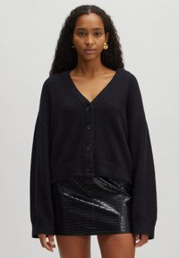 Zwarte gebreide cardigan met een v-hals, oversized pasvorm en vijf knopen aan de voorkant. Draagt over een glanzende zwarte rok met krokodillenpatroon.