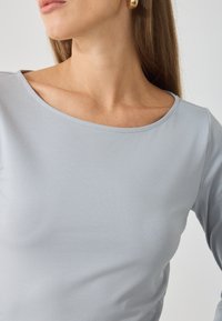 Maglietta a maniche lunghe grigio chiaro con una scollatura ampia e arrotondata. Tessuto liscio e dettagli di cucitura sottili sui bordi. Accentuata da grandi orecchini dorati.