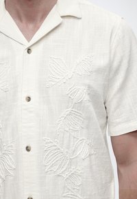 Λευκό πουκάμισο με κοντά μανίκια και κουμπιά, με ανάγλυφη κεντημένη floral διακόσμηση και γιακά τύπου καμπ, που φοριέται από άτομο με ορατό λαιμό και χέρι.