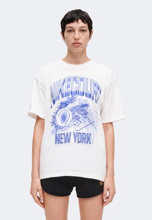 Personne aux cheveux courts et foncés porte un t-shirt blanc Nike Court New York avec un motif de tennis bleu et un short noir sur un fond clair uni.