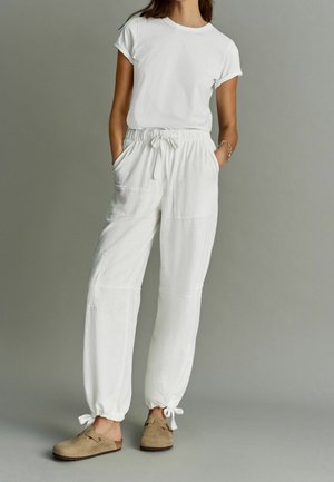 Kvinde iført hvid t-shirt med korte ærmer og hvide bukser med snøre i taljen og sidelommer, kombineret med brune slip-on sko mod en grå baggrund.