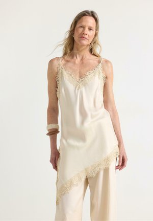 Donna bionda che indossa un abito in seta color crema con pizzo in rifinitura sopra pantaloni larghi abbinati, accessoriata con due braccialetti sul polso sinistro.