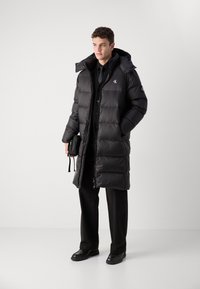 Schwarze Puffjacke mit durchgehendem Reißverschluss, Kapuze und Logodetail. Getragen über einem schwarzen Hemd mit Knöpfen und einer Hose, dazu eine kleine Umhängetasche.