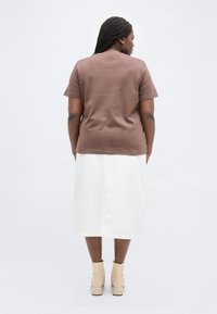 Γυναίκα με πλεξούδες, φορεμένη με καφέ μπλούζα κοντού μανικιού, λευκή φούστα μέσης μήκους και μπεζ μπότες αστραγάλου, στέκεται γυρισμένη προς τα πίσω σε λευκό φόντο.