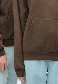 Brauner Fleece-Kapuzenpullover mit einer Vorderentasche, gerippten Bündchen und Saum, kombiniert mit hellblauen Jeanshosen. Fokus auf Textur und Farbkontrast.