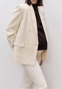 Blazer beige doppiopetto con rever a intaglio, abbinato a una maglia scura e pantaloni bianchi. Tessuto morbido con dettagli di bottoni.