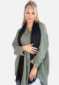Chemise olive, à boutons, en tissu texturé, portée sous une écharpe noire avec des bords effilochés. Les accessoires incluent des bagues en or.