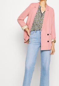 Blazer rosa con un design doppiopetto, maniche a fantasia beige, indossato sopra una blusa floreale e jeans azzurri chiari.