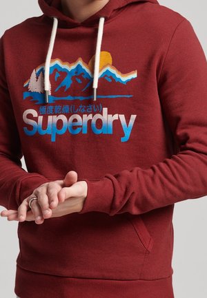 Personne portant un sweat à capuche rouge Superdry avec un motif montagne et soleil, tenant sa main gauche avec sa main droite devant un fond gris.