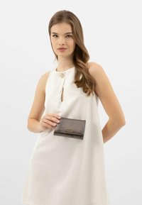 Portafoglio grigio texture, tenuto contro un vestito bianco, con un piccolo logo dorato e una chiusura a patta con un delicato motivo in rilievo.