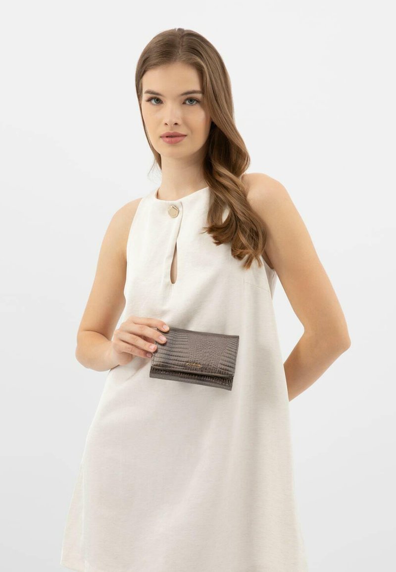 Portafoglio grigio texture, tenuto contro un vestito bianco, con un piccolo logo dorato e una chiusura a patta con un delicato motivo in rilievo.